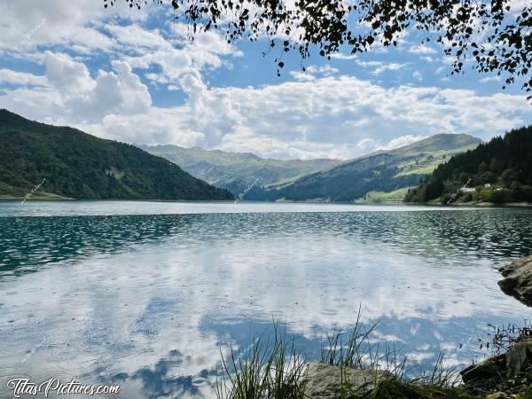 Photo Lac de Roselend : Le Lac de Roselend sous la pluie, mais avec du soleil en même temps. Résultat : sa belle couleur turquoise disparaît sous les gouttes mais on voit encore le reflet des nuages dans l’eau. Ça crée une drôle d’ambiance 🤭😅c, Tita’s Pictures, Les Alpes, Lac de Roselend, Beaufortin, Savoie